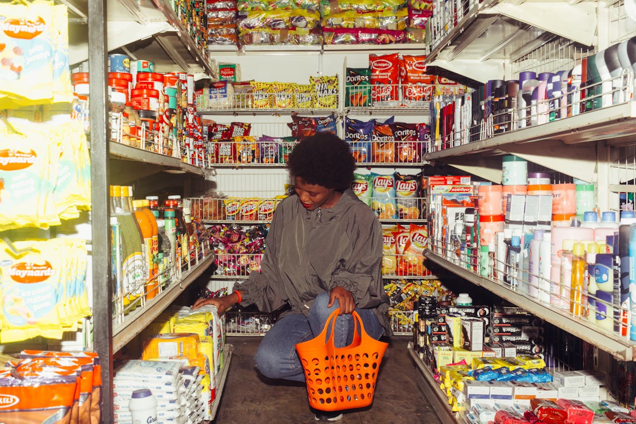 Full length of adult ethnic female buyer selecting goods while squatting with shopping bag among grocery shelves